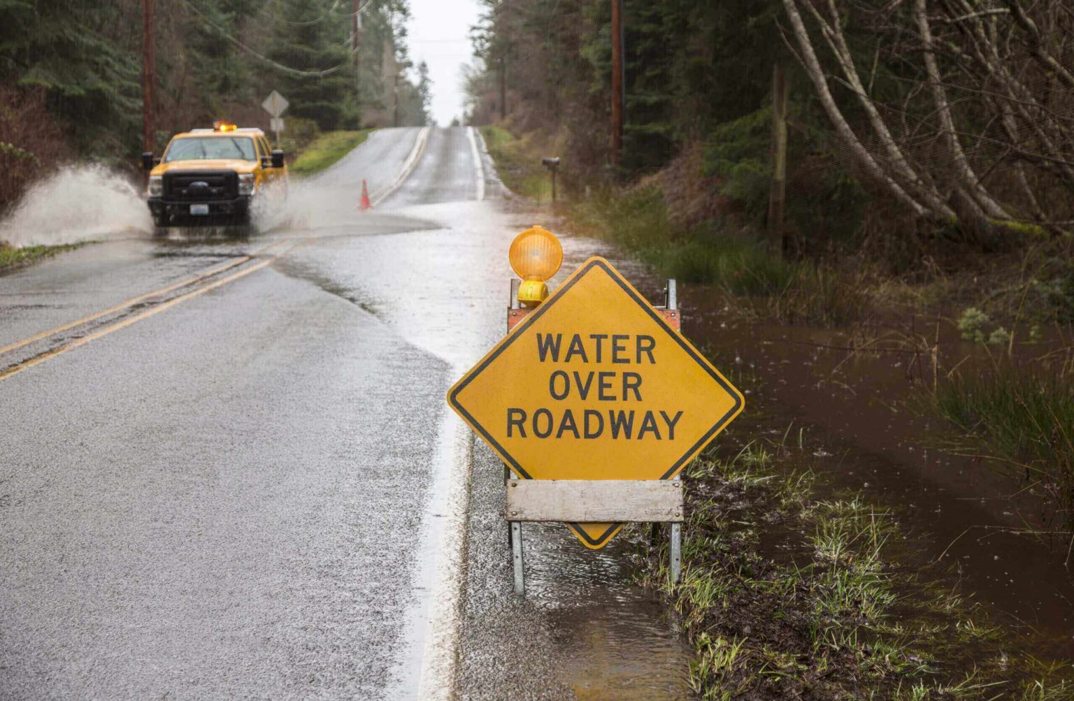 Emergency vehicle driving through stormwater on a road flooded by a natural disaster rain storm. A warning sign indicates a hazard.